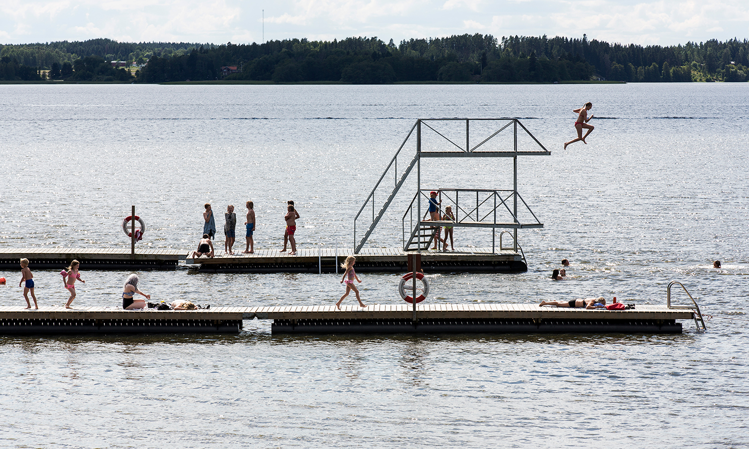 Foto på Bredsandsbadet och barn som hoppar från brygga och hopptorn ner i vattnet.