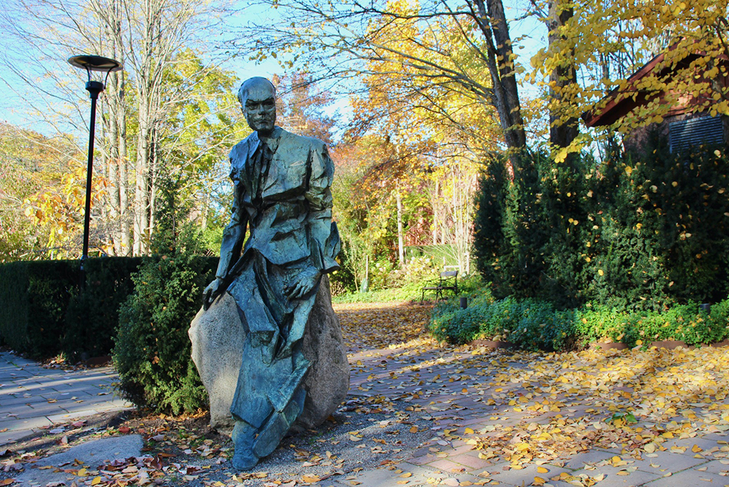 Foto på en staty utanför Fridegårdsparkens ingång. Statyn föreställer Jan Fridegård som sitter lutad mot en sten. Bakom statyn syns ingången till parken.