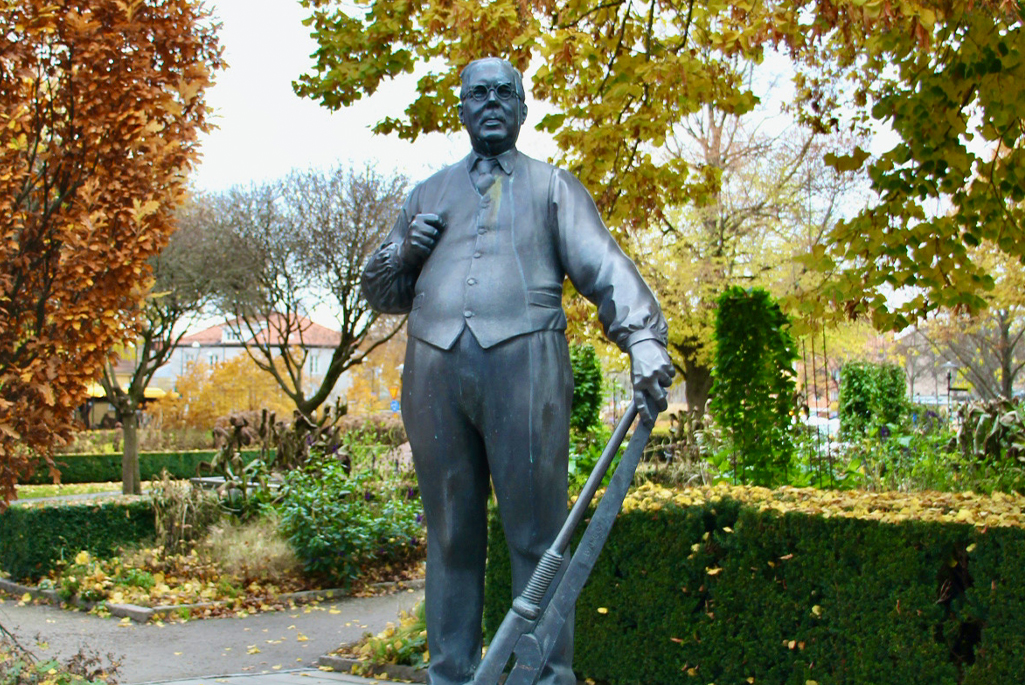 Foto på en staty som står utanför Gustav Adolfs plan. Statyn föreställer JP Johansson som håller i ett stort verktyg. Bakom statyn syns parken.